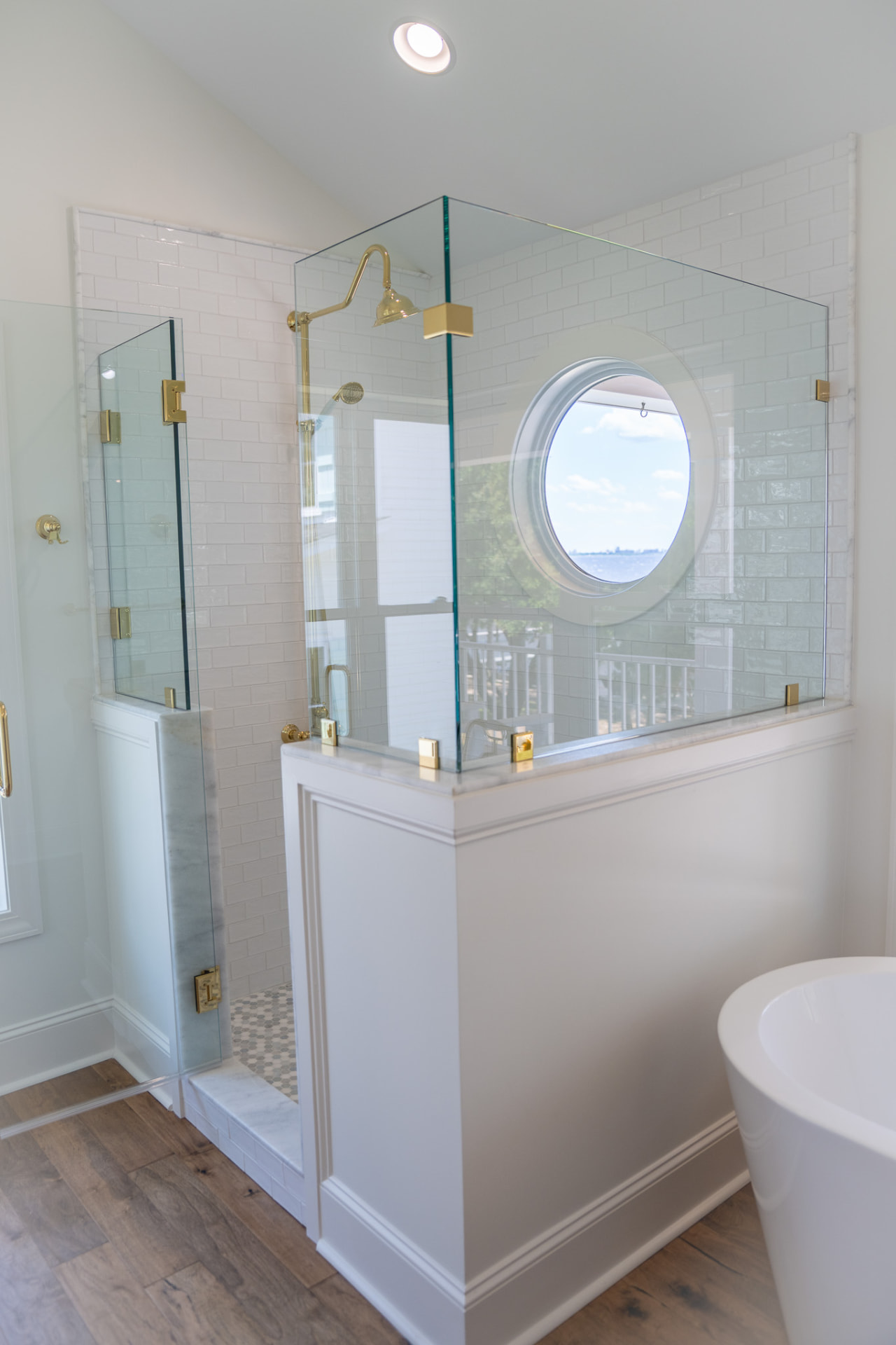 Primary Bath — Porthole Shower — Frameless glass enclosure over a marble knee wall, subway tile surround and a porthole window framing the water.