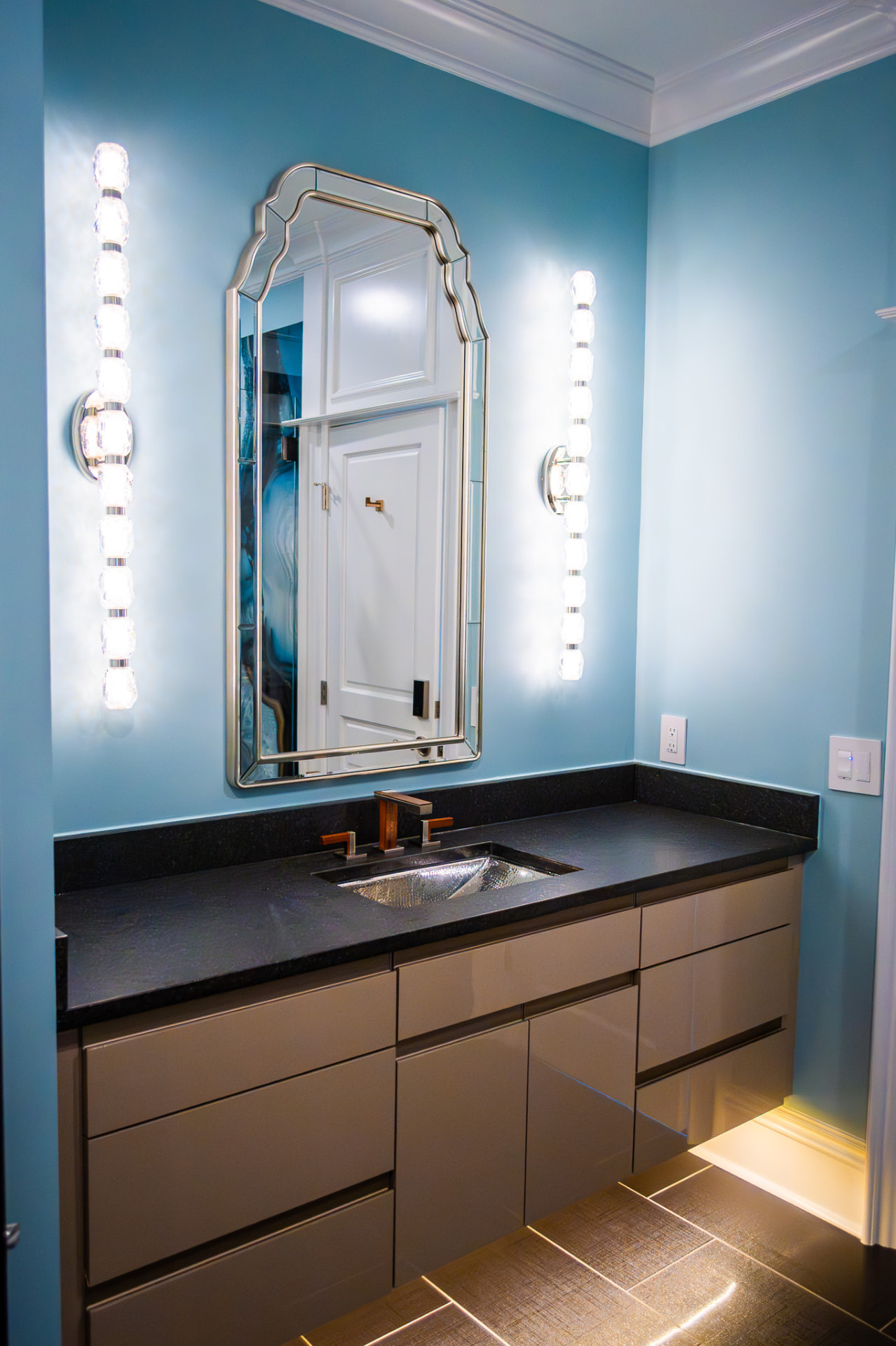 Vanity & Sconce Detail — Honed black stone counter and a hammered metal basin set against soft blue walls, flanked by crystal sconces and toe-kick lighting.