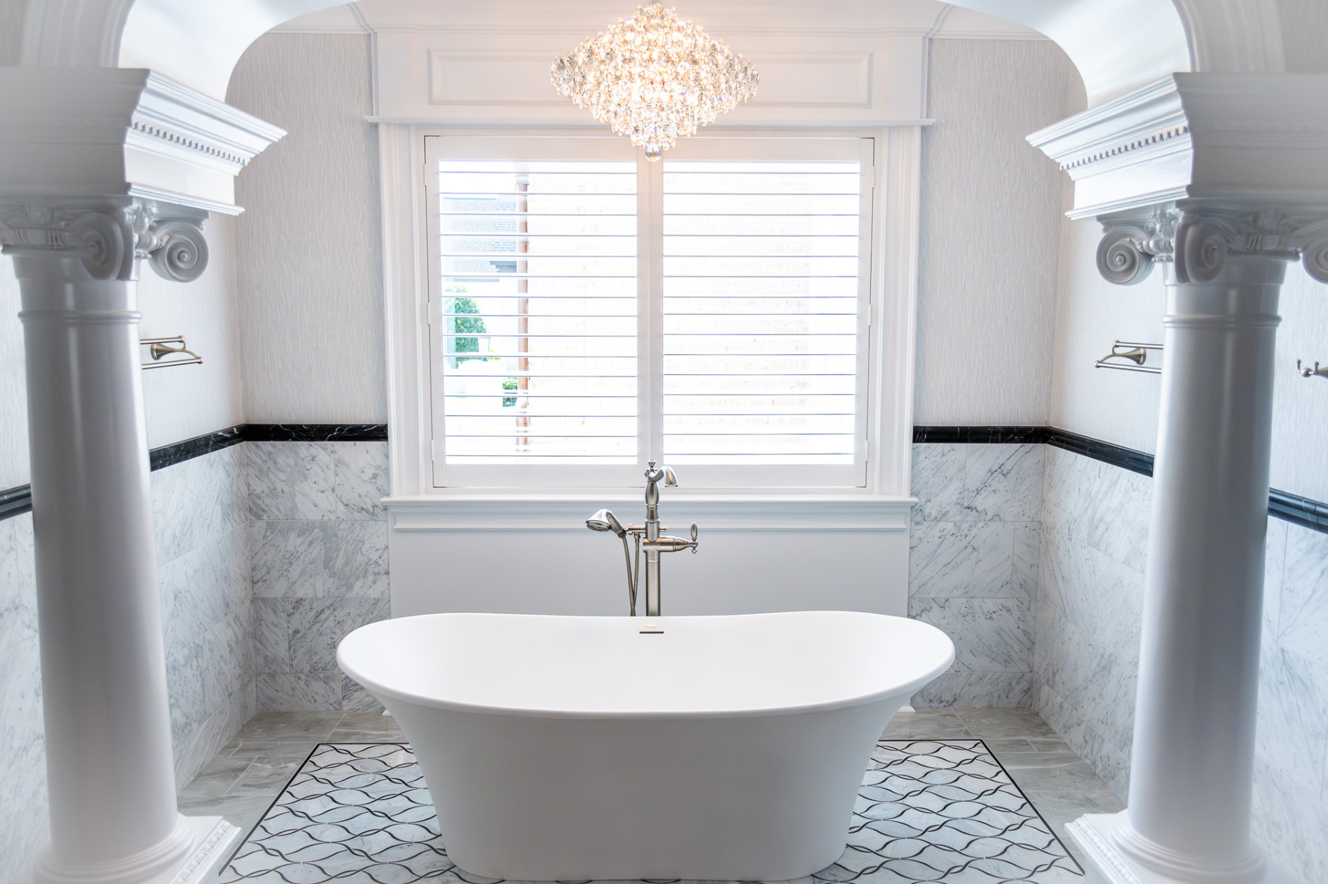 Primary Bath — Chandelier — Freestanding tub set beneath a crystal chandelier, with marble-clad walls, classical columns and polished stone flooring.