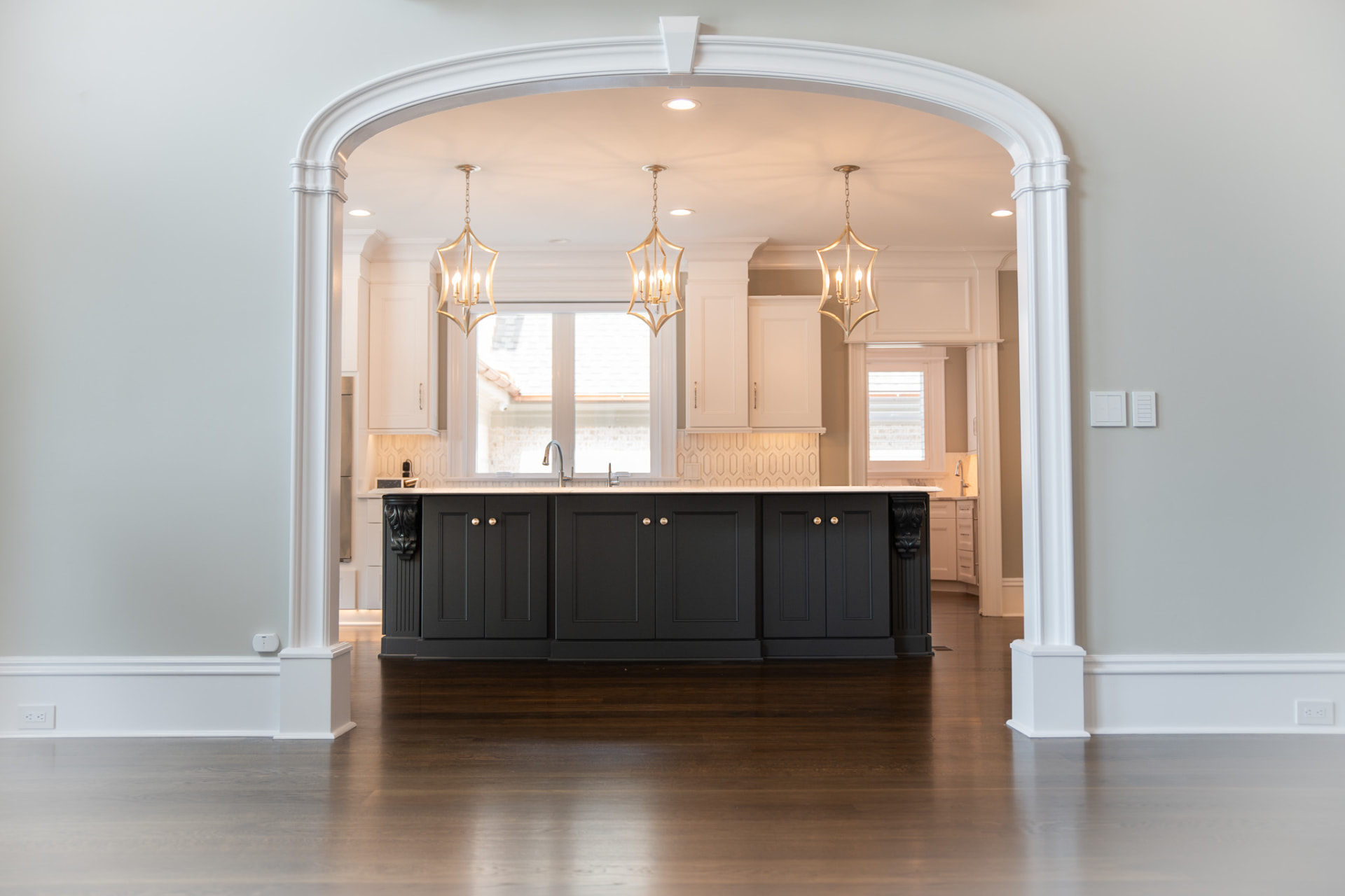 Kitchen Through Arched Opening — Black island with brass hardware and pendant lights, framed by a deep cased archway and site-finished oak flooring.