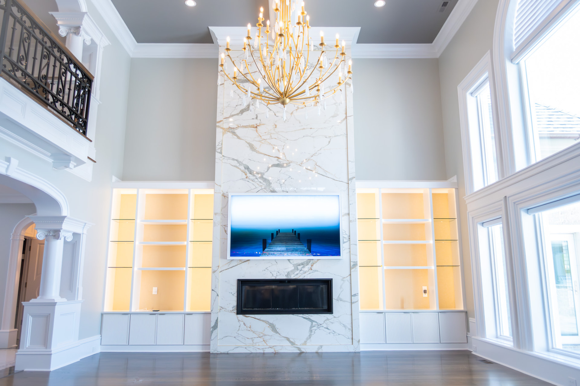 Great Room & Stair Hall — Double-height living volume with a marble-clad fireplace wall, custom built-ins and a wrought-iron balcony rail.