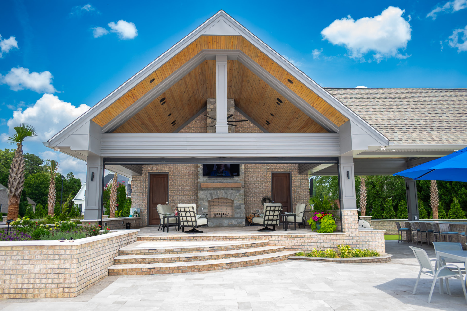 Custom outdoor pavilion with a high gable roof, stone fireplace and brick patio under a bright blue sky
