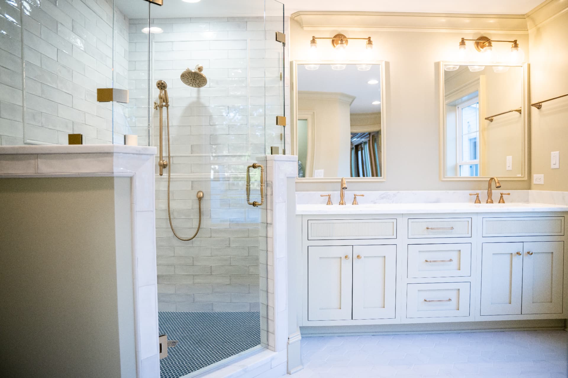 Primary Suite Bath — Double vanity with fluted drawer fronts, brushed brass fittings and a glass shower with penny-round tile floor.