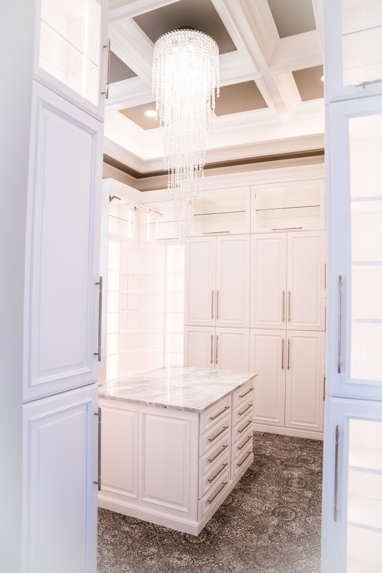 Dressing Room — Coffered ceiling, glass-front cabinetry and a marble-topped island centred beneath a crystal fixture.
